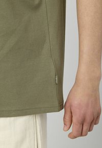 T-shirt en coton vert olive avec un col rond, des manches courtes et une petite étiquette logo sur le côté inférieur. Texture lisse, coupe décontractée.