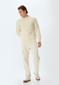 Cremfarbener, gerippter Strickpullover mit Rundhalsausschnitt, kombiniert mit hellbeigen Cargo-Hosen mit Seitentaschen und tanfarbenen Sneakers.