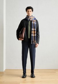 Hnedá kožená taška cez rameno, sivá bunda, zelený sveter, námornícke nohavice a modro-oranžový károvaný šál, vystavené na modeli na jednoliatom pozadí.