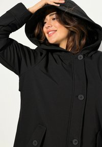 Cappotto nero con cappuccio, maniche lunghe, chiusura frontale con bottoni e tasche laterali. Il tessuto ha una texture liscia e un design dalla vestibilità rilassata.