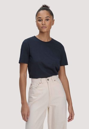 Frau mit hochgestecktem Haar, die ein einfaches dunkelblaues T-Shirt trägt, das in eine hoch taillierte, hellbeige Hose mit weitem Bein gesteckt ist, steht vor einem neutralen Hintergrund.