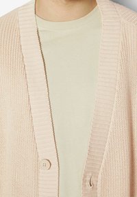 Gilet en tricot rose pâle avec un col côtelé et un plastron avant, doté d'une fermeture à un bouton. Texture douce sur une chemise beige.