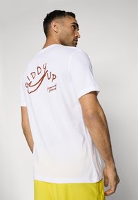 Hvit bomulls t-skjorte med en brun grafisk trykk på ryggen som sier "Giddy Up" med et swoosh-design. Rund hals og korte ermer.