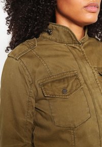 Olivgrüne Jacke mit hohem Kragen, zwei vorderen Taschen und Knopfverschlüssen. Hergestellt aus robustem Stoff mit strukturierter Oberfläche.