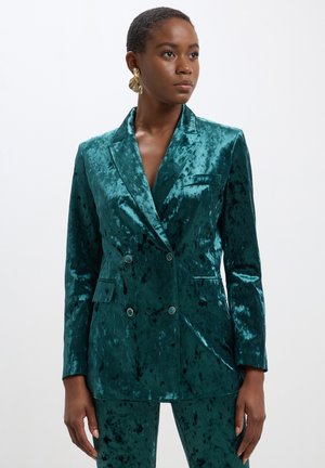 Blazer doppiopetto in velluto teal con rever a punta, due tasche frontali e bottoni dorati. Tessuto liscio con una leggera lucentezza.