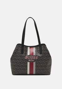 Kangaskassi, jossa on harmaasta kuvioidusta materiaalista valmistetut mustat kahvat. SISÄLTÄÄ pystysuorat punaiset ja kerma-valkoiset raidat sekä "GUESS EST.1981" -logo.