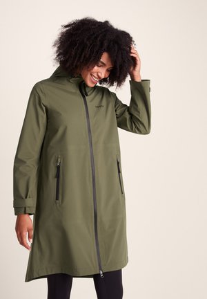 Woman with curly hair wearing a long olive green raincoat with black zippers, smiling and adjusting hair against a plain background.