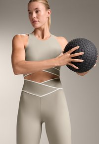 Bežna športna majica z izrezom in belimi detajli, v kombinaciji z ujemajočimi se leggins hlačami z vzorcem križa. Drži teksturirano črno medicinsko žogo.