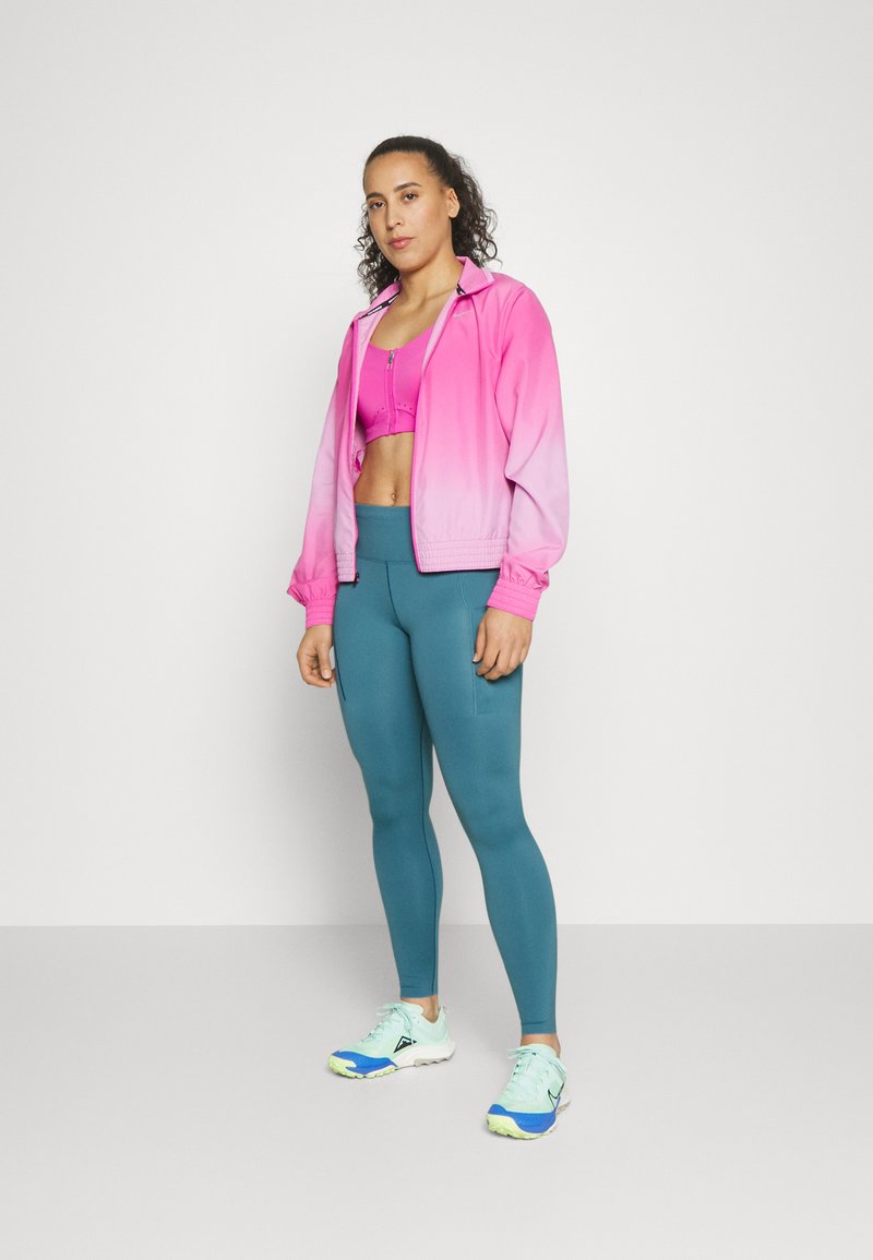 Pink-gradient Sportjacke über einem passenden Crop-Top, kombiniert mit türkisfarbenen Leggings und mehrfarbigen Laufschuhen. Glatter, elastischer Stoff.