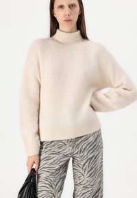 Helles Beige, hochgeschlossenes Sweater aus weichem, strukturiertem Strick. Kombiniert mit grauen Hosen mit einem schwarzen Zebramuster. Das Modell hält eine schwarze Tasche.