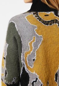 Gestrickter Pullover mit hohem schwarzem Kragen, strukturiertem Wollstoff in Grau, Grün und Senfgelb mit abstrakten Mustern und kontrastierenden Nahtdetails.