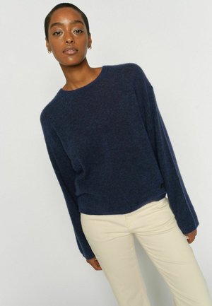 Frau in einem marineblauen Langarmpullover und hellbeigen Hosen, stehend vor einem schlichten hellen Hintergrund.