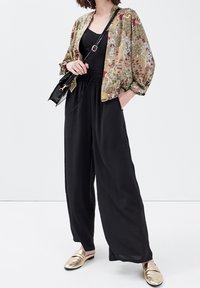 Veste kimono légère à motif floral en olive et or, portée avec une combinaison noire et des mocassins métalliques dorés. Sac à bandoulière noir.