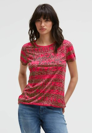 Woman with dark hair wearing a red and brown patterned short-sleeve shirt with text and blue jeans, standing with hands in pockets.