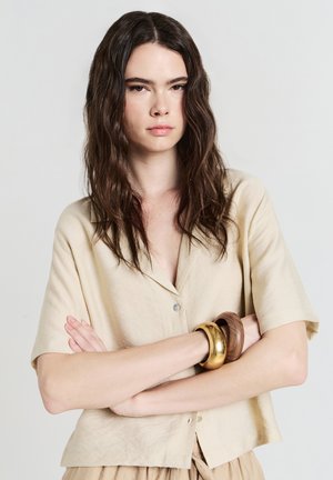 Giovane donna con capelli castani mossi indossa una maglietta beige a maniche corte e grandi braccialetti in legno e oro, in piedi con le braccia incrociate.
