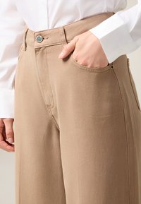 Pantalones de pierna ancha beige con una textura suave, un bolsillo frontal y un cierre de botón metálico en la cintura. Costuras delgadas acentúan el diseño.