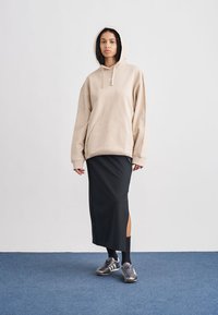 Μπεζ φαρδιά φούτερ με τσέπη μπροστά, σε συνδυασμό με μαύρη φούστα μήκους μέχρι το γόνατο με πλαϊνές σχισμές και γκρι αθλητικά παπούτσια.