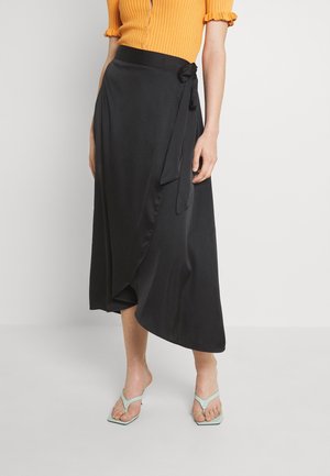 Femme portant une jupe portefeuille noire asymétrique nouée à la taille, un haut côtelé orange à manches courtes et des sandales bleu clair ouvertes à l’avant.