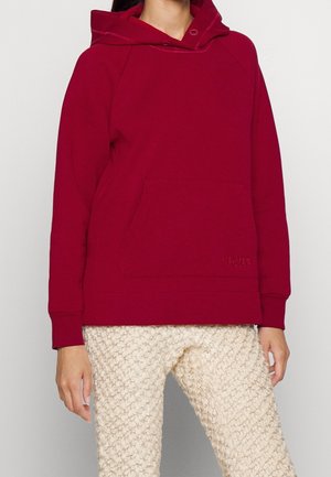 Person wearing a red hooded sweatshirt with front pocket and textured cream-colored pants, standing against a plain background.