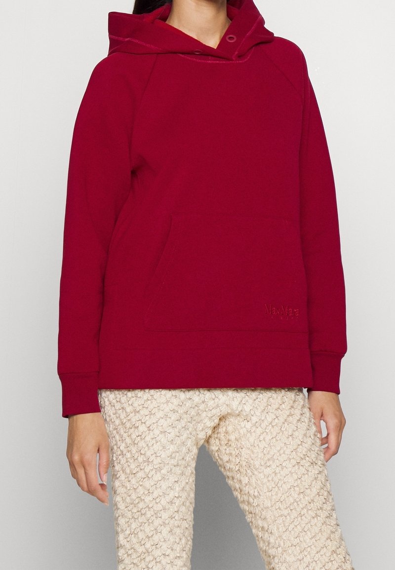 Personne portant un sweat à capuche rouge avec poche avant et pantalon texturé de couleur crème, debout devant un fond uni.