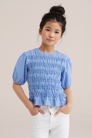 Fiatal lány sötét hajjal, kontyban, texturált világoskék, puffos ujjú felsőt és fehér nadrágot visel, kezét a zsebében tartva, semleges háttér előtt áll.