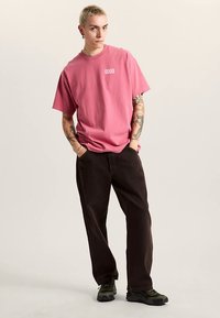 Roze katoenen T-shirt met de tekst "GOOD" op de borst, gecombineerd met donkerbruine broek. Gestructureerde sneakers met een groene accent.