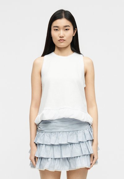 Femme aux longs cheveux noirs, portant un haut blanc sans manches et une mini-jupe à volants à étages rayée bleu et blanc, debout face à l'avant.