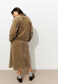 Veste en velours côtelé marron et jupe midi assortie, arborant une texture côtelée et une coupe décontractée. Les chaussures sont marron avec des chaussettes grises.