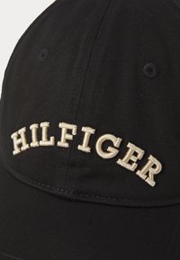 Svart baseballcap laget av bomullstoff, med hvit brodert tekst "HILFIGER" på forsiden. Bøyd skjerm med synlige sømdetaljer.