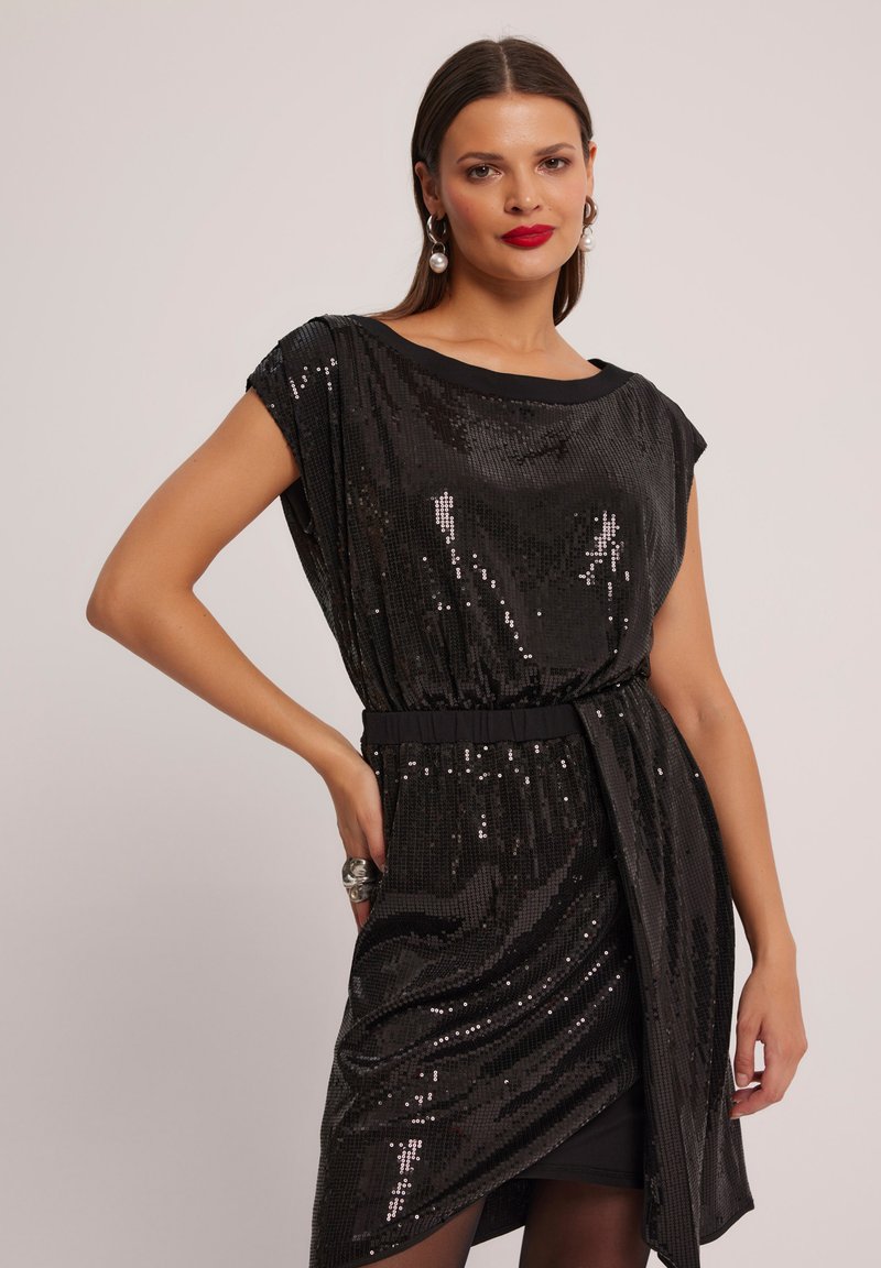 Czarna sukienka z cekinami o luźnym kroju, krótkich rękawach i podkreślonej talii. Ma okrągły dekolt i asymetryczny dół.