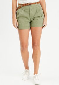 Shorts vert olive avec taille haute, plis et ourlets retroussés. Portés avec une ceinture tressée marron et associés à des baskets blanches.