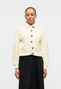 Γυναίκα με πλεξούδες στα μαλλιά, φοράει ένα κρεμ πλεκτό ζακέτα με κουμπιά πάνω από ένα λευκό πουκάμισο με γιακά και μια μαύρη φούστα, με φόντο απλό λευκό.