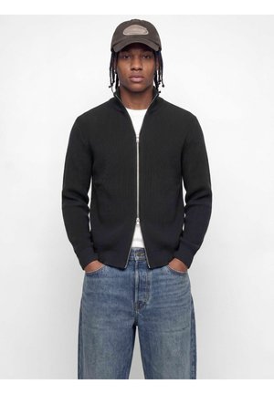 Homme aux cheveux tressés portant un pull zippé foncé, un jean bleu, une chemise blanche et une casquette de baseball sombre sur un fond uni.