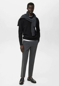 Schwarzer Pullover, grauer Schal über den Schultern, weißes T-Shirt darunter, schmale graue Hose und schwarze elegante Schuhe. Schlichtes, modernes Design.