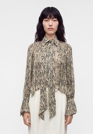 Frau mit langen dunklen Haaren, die eine beige-schwarze Bluse mit Schlangenprint, einer gebundenen Schleife am Kragen und ausgestellten Ärmeln trägt, kombiniert mit weißen Hosen.