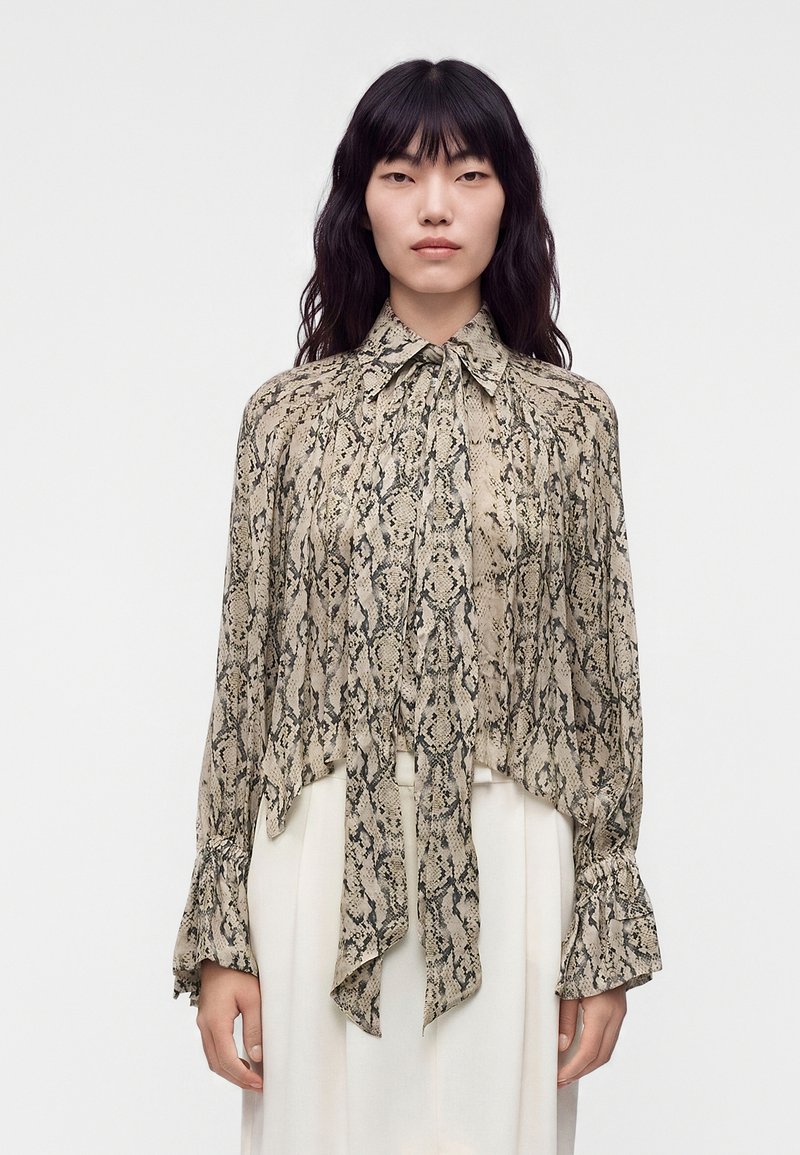 Woman with long dark hair wearing a beige and black snake print blouse with a tied neck bow and flared sleeves, paired with white pants.