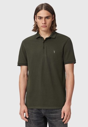 Ung mand med mellembrunt hår iført en mørkegrøn kortærmet polo-shirt og mørke jeans, stående foran en ensfarvet baggrund.
