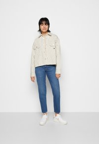 Striped, button-up shirt in cream and black with two chest pockets, paired with fitted blue jeans and white sneakers.