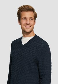 Marineblauer Pullover mit V-Ausschnitt-Design, das ein strukturiertes Rautenmuster aufweist. Hergestellt aus Strickmaterial mit gerippten Bündchen.