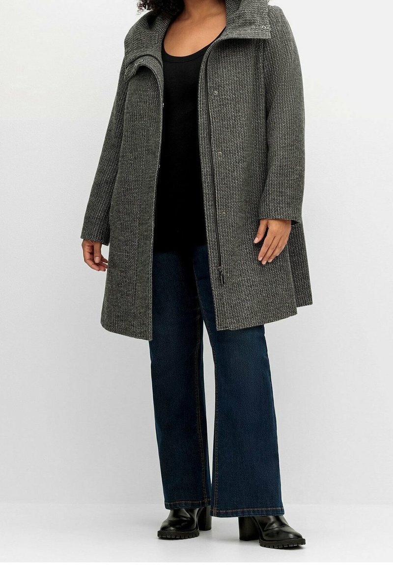Grijze geweven jas met een hoge kraag en een rits aan de voorkant. Gecombineerd met een zwarte top en blauwe flared jeans, aangevuld met zwarte enkellaarsjes.