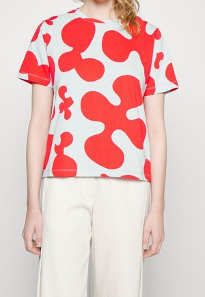 Kobieta ubrana w biały T-shirt z dużymi nieregularnymi czerwonymi kształtami i luźne białe spodnie, stojąca na tle jednolitego jasnego tła.