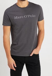 Szara bawełniana koszulka z krótkimi rękawami i okrągłym dekoltem, z nadrukowanym logo "Marc O’Polo" w jaśniejszym odcieniu szarości na klatce piersiowej.