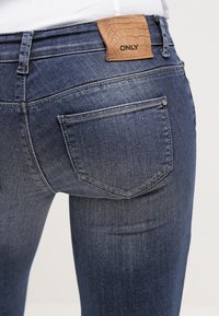 Jeans de denim azul oscuro con un diseño ajustado, que cuentan con bolsillos traseros y una etiqueta de imitación cuero marrón con "ONLY" grabado.