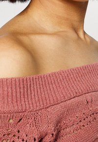 Różowy, dziergany sweter z dekoltem typu off-the-shoulder, z ribbowanym wykończeniem i ażurowym wzorem wzdłuż dołu. Miękka tekstura.
