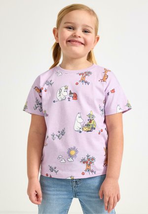 Hymyilevä nuori tyttö, jolla on vaaleat lettinutturat ja joka pukeutunut liilanväriseen T-paitaan, jossa on värikkäitä Muumi-hahmoja, sekä vaaleansiniset farkut.