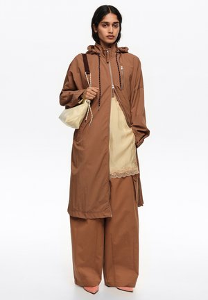 Mujer con abrigo marrón con capucha, pantalones de pierna ancha, delantal crema con encaje, sosteniendo un bolso beige de hombro, de pie frente a fondo blanco.