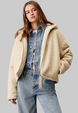 Romige faux fur jas met een capuchon, ritsluiting en brede mouwen, gelaagd over een blauwe denimshirt en high-waisted denim jeans. Label zichtbaar.
