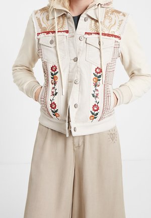 Cream hooded denim jacket with floral embroidery on front panels worn with beige wide-leg pants. Hands in pockets.