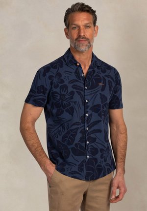 Man van middelbare leeftijd met een zout-en-peper baard, gekleed in een marineblauw shirt met korte mouwen en bloemenpatronen, en beige broek, staand met één hand in de zak.