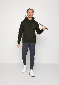 Homem vestindo um capuz preto e calças de treino azuis marinho, segurando um taco de golfe ao ombro, em pé contra um fundo claro e simples.